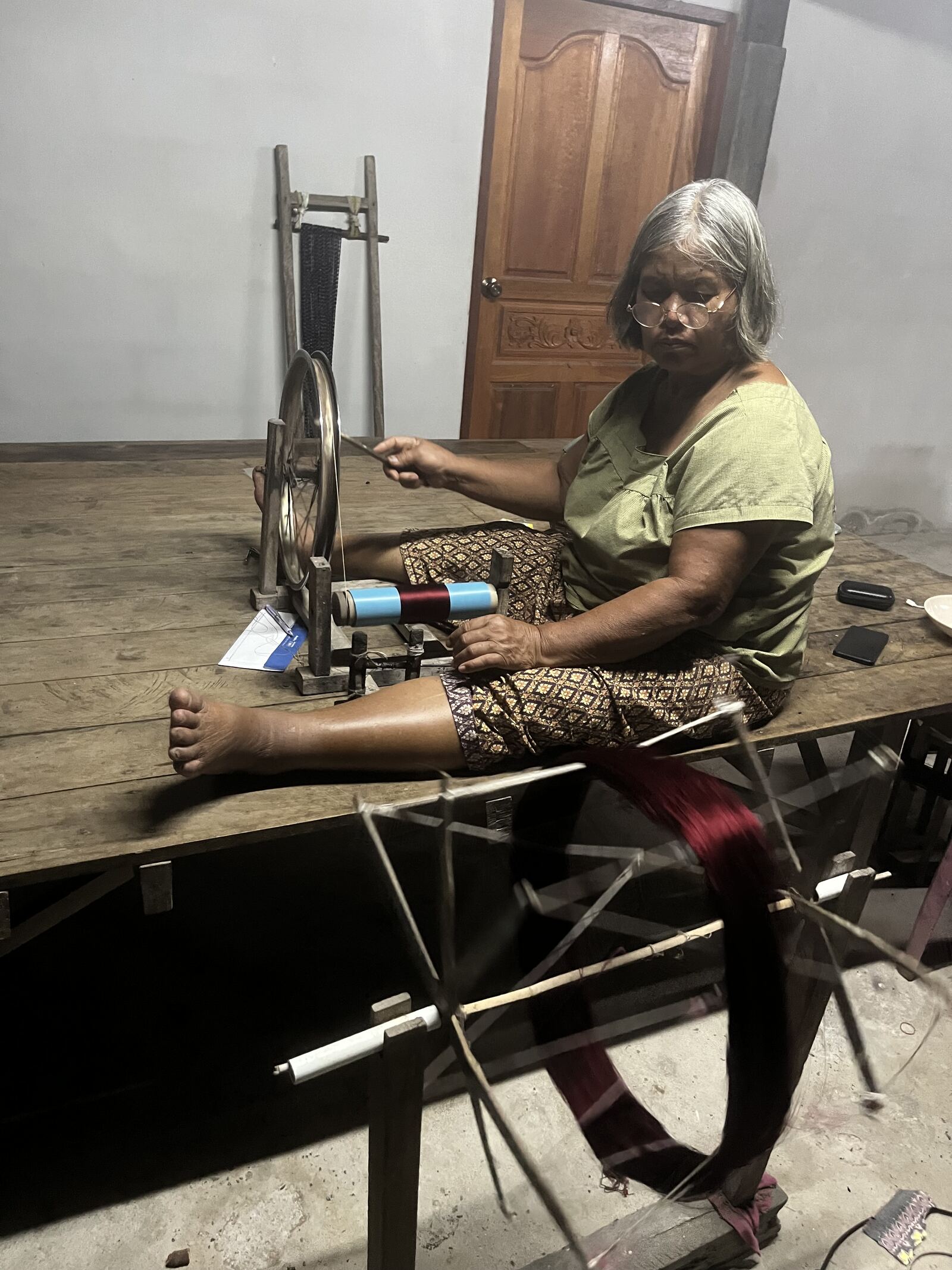 Wilai at the spinning wheel, winding deep red silk thread onto a turning frame